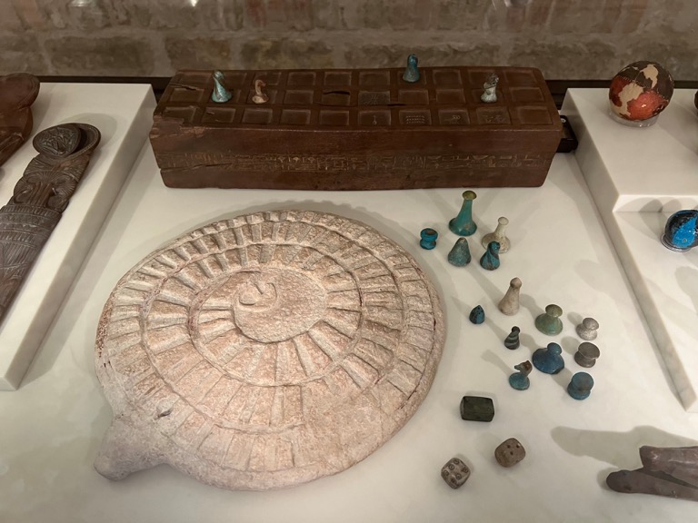 <a href="https://recherche.smb.museum/detail/779568/senet-spielbrett-des-sennefer?language=de&question=senat+game&limit=15&sort=relevance&controls=none&collectionKey=AMP*&objIdx=0">Senet Game</a> in the back middle